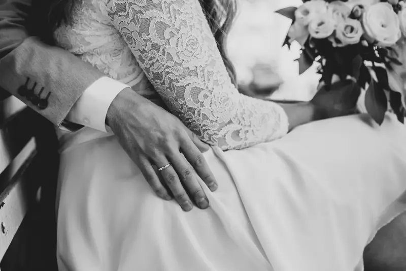 Seine Hand auf ihrem Spitzenkleid, daneben der Brautstrauß, in Schwarzweiß